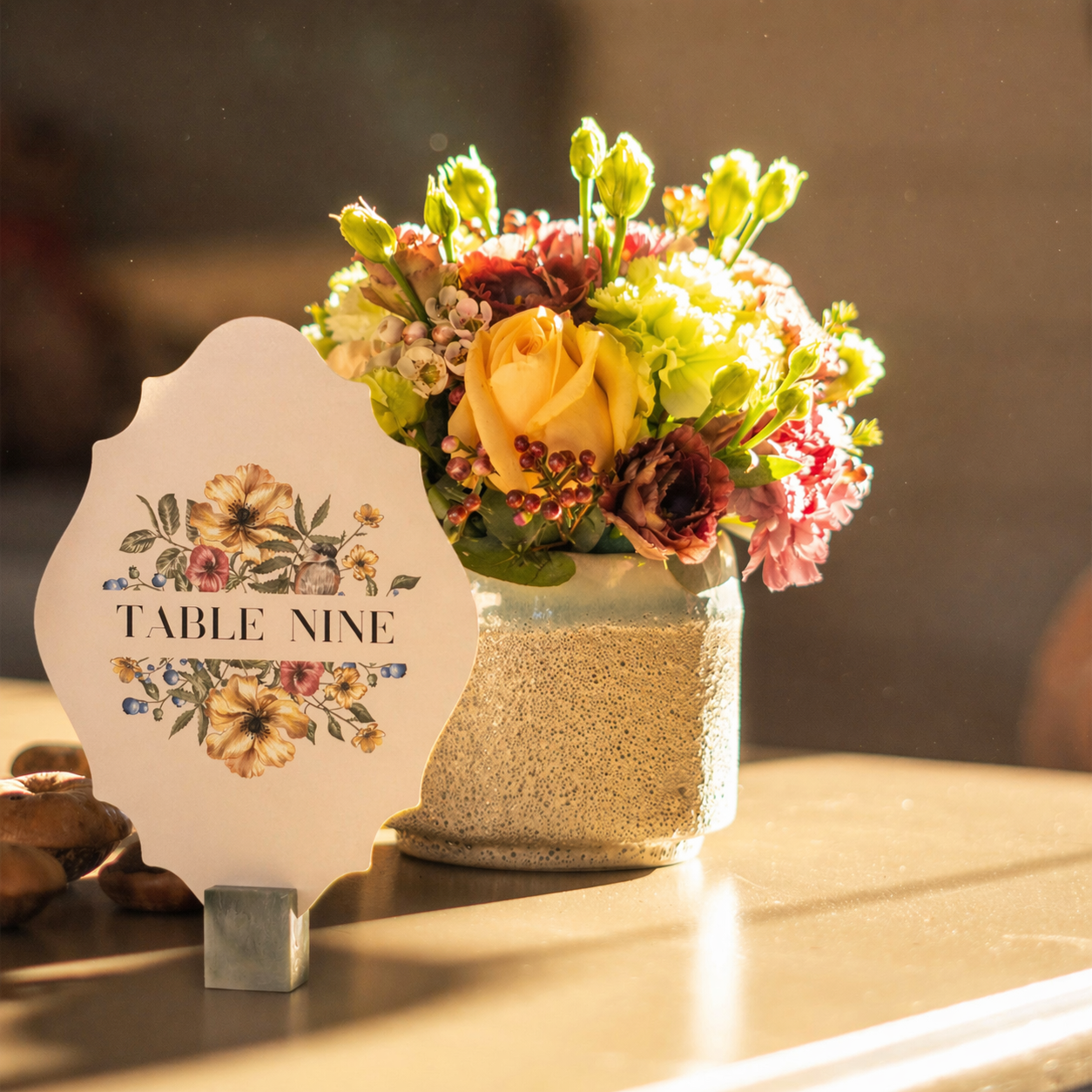 Floral arrangement in a textured vase with a  die-cut table number sign on a wooden surface.