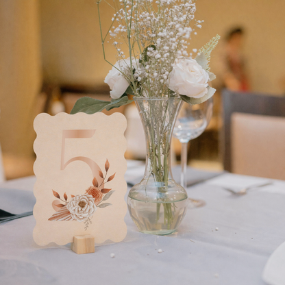 Wave shape table number card with floral design next to a vase with white flowers on a table.