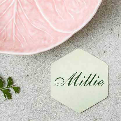 Hexagonal green name card with 'Millie' on a textured surface with a pink ceramic dish.