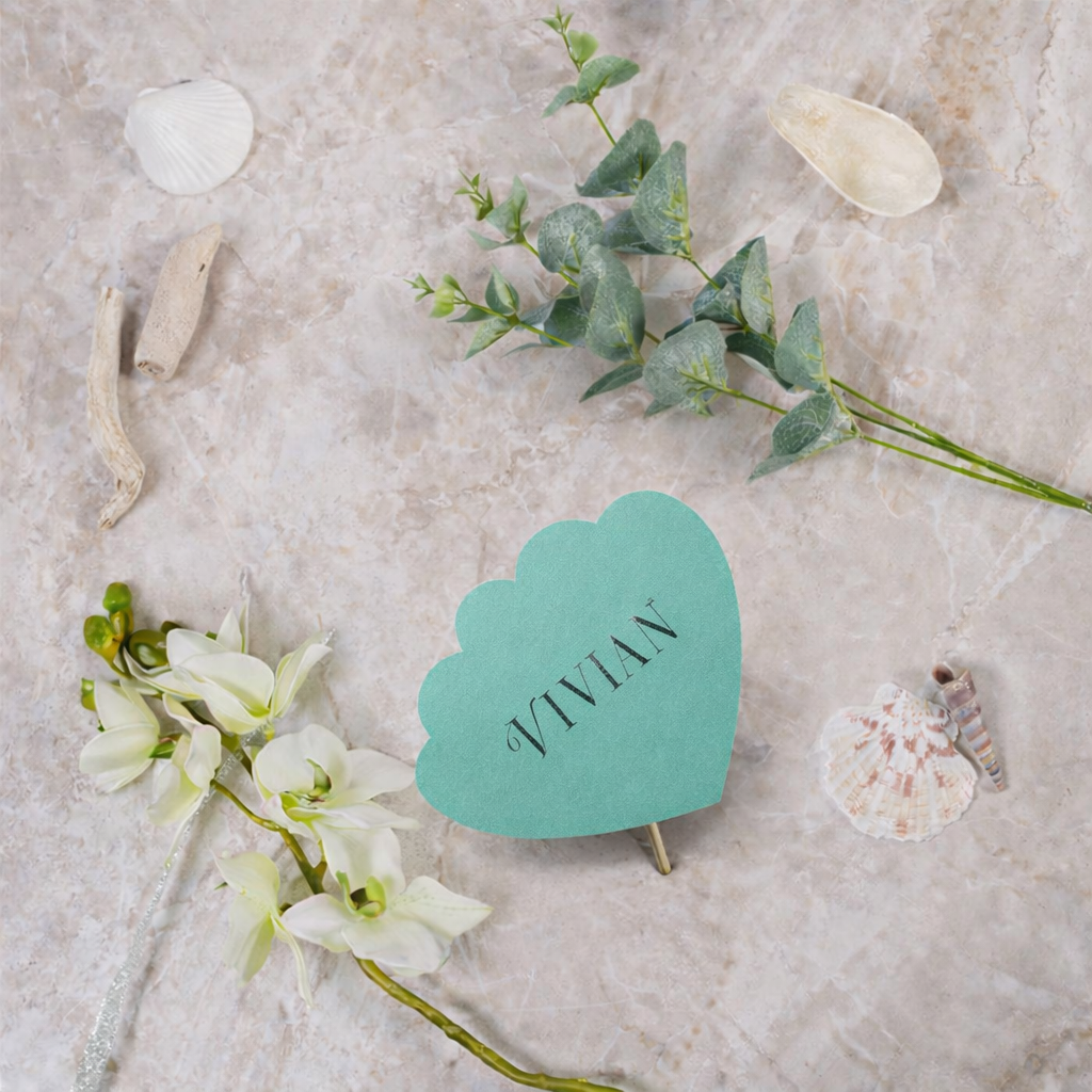 Turquoise shell shaped escort card with 'Vivian' on a stone surface with flowers and shells